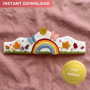 Puede incluir: Una corona de fieltro blanco con un arcoíris, mariposas, sol y hojas de otoño. La corona tiene un diseño caprichoso con un arcoíris arqueado sobre una base verde. El texto "INSTANT DOWNLOAD" está en la parte superior.
