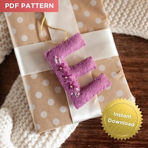 May include: A handcrafted, lavender felt letter "E" ornament, embellished with small floral and bead accents. The ornament hangs from a gold cord and rests on a polka-dot wrapped gift with a white ribbon. The words "PDF Pattern" and "Instant Download" are visible.
