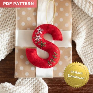 Puede incluir: Adorno navideño con forma de letra "S" de fieltro rojo con bordado floral, colgado de un cordón dorado. El adorno está sobre un regalo envuelto con una cinta blanca. Se ven las palabras "PDF PATTERN" y "Instant Download".