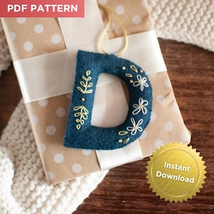 May include: A dark teal felt letter "D" ornament with gold floral embroidery and white flower accents. The ornament hangs from a gold cord and is displayed on a wrapped gift. The words "PDF PATTERN" and "Instant Download" are visible.