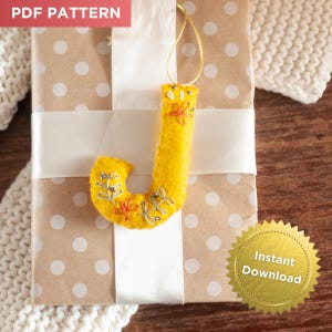 May include: A yellow felt letter "J" Christmas ornament with floral embroidery and silver bead accents. The ornament hangs from a gold string and is placed on a wrapped gift with a white ribbon. The words "PDF PATTERN" and "Instant Download" are visible.
