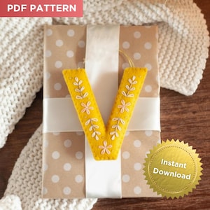 May include: A yellow felt letter "V" ornament with floral embroidery, placed on a wrapped gift with a white ribbon. The gift is wrapped in brown paper with white polka dots. The text "PDF PATTERN" is in the top left corner.