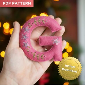 May include: A pink felt letter "G" with floral embroidery. The letter is held in a hand, with the text "PDF PATTERN" in the upper left corner and a gold "Instant Download" seal in the lower right corner.