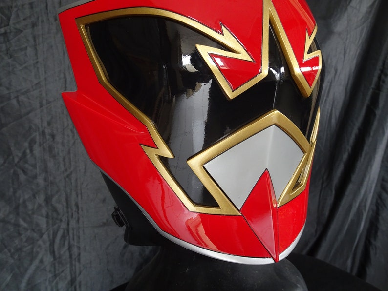 Op de afbeelding: Rode, zwarte en gouden plastic helm met een gestileerd ontwerp. De helm heeft een zwart vizier en een gouden rand.