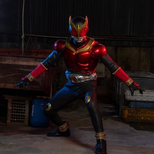 May include: A person in a red and black superhero costume with gold accents, including a helmet, chest armor, and belt. The costume has a metallic sheen and is posed in a dynamic stance. The background is an industrial setting with concrete and metal.