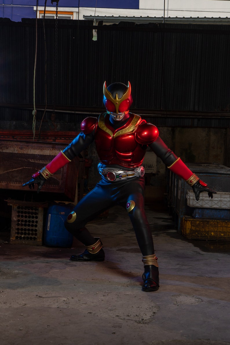May include: A person in a red and black superhero costume with gold accents, including a helmet, chest armor, and belt. The costume has a metallic sheen and is posed in a dynamic stance. The background is an industrial setting with concrete and metal.