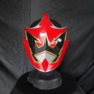 Op de afbeelding: Rode, zwarte en gouden Power Rangers helm met een stervormig ontwerp. De helm is gemaakt van plastic en heeft een glanzende afwerking.