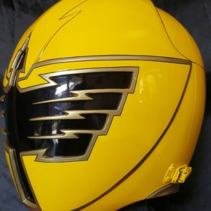 Op de afbeelding: Gele plastic helm met een zwart en gouden ontwerp. De helm heeft een gouden rand rond de randen en een zwart vizier.