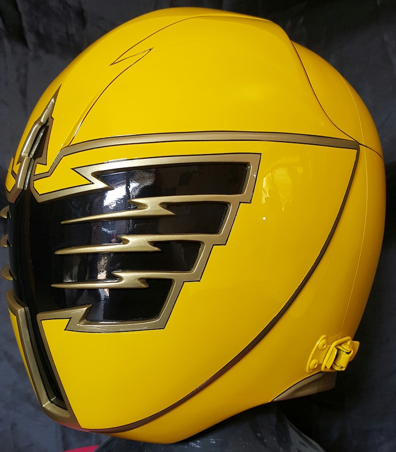 Op de afbeelding: Gele plastic helm met een zwart en gouden ontwerp. De helm heeft een gouden rand rond de randen en een zwart vizier.