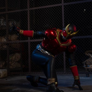 May include: A person in a red and black superhero costume, kneeling on a concrete surface. The costume features a helmet with gold accents, red armor, and black pants. The background includes a chain-link fence and a dark, industrial setting.