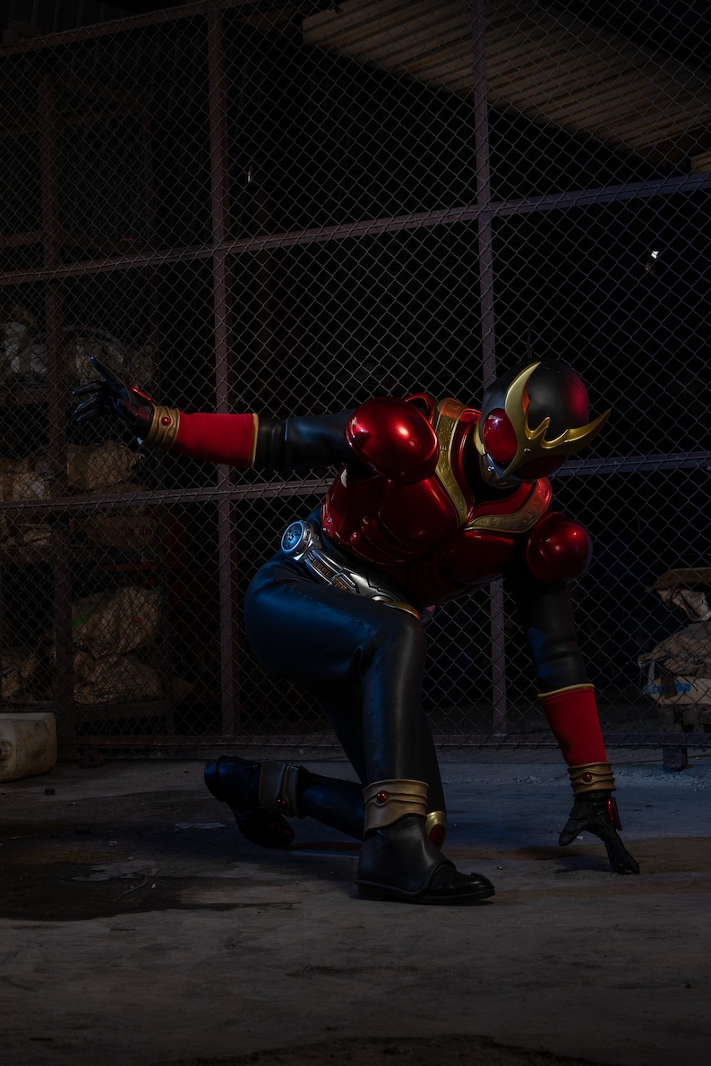May include: A person in a red and black superhero costume, kneeling on a concrete surface. The costume features a helmet with gold accents, red armor, and black pants. The background includes a chain-link fence and a dark, industrial setting.