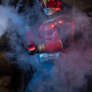 May include: A red and gold superhero costume with a helmet, chest armor, and arm gauntlets. The costume features a red visor and gold accents. The figure is surrounded by blue and white smoke.