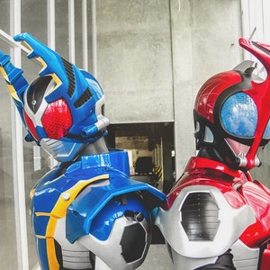 May include: Two detailed cosplay helmets, one blue and one red, stand side-by-side. The blue helmet has a distinctive design with yellow accents and a red visor. The red helmet features a sleek, streamlined shape with a blue visor. Both are part of a cosplay theme.