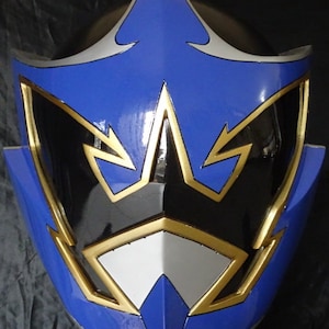Op de afbeelding: Een blauwe en zwarte helm met gouden accenten. De helm heeft een scherp, hoekig ontwerp met een puntige bovenkant.