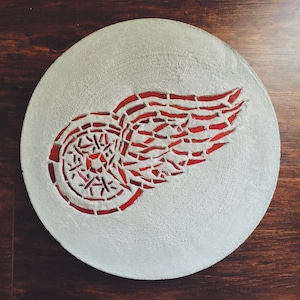 May include: A round, white, concrete coaster with a red and white mosaic design of a hockey team's logo. The logo features a red wing with a white circle in the center.