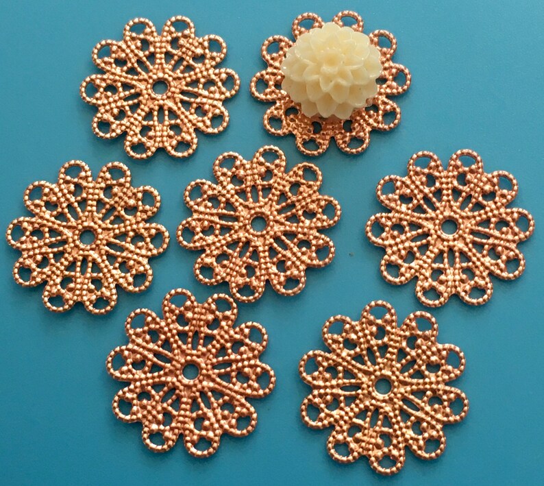 May include: Six rose gold metal filigree embellishments with a circular design. The embellishments have a delicate, intricate pattern and a hole in the center.