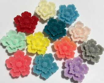 Könnte beinhalten: Sortiment von 12 bunten, blumenförmigen Harz-Cabochons. Die Cabochons sind in verschiedenen Farben, darunter Rot, Gelb, Blau, Grün, Rosa und Grau. Jeder Cabochon hat ein erhabenes Blumendesign.