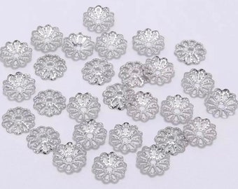 May include: A collection of silver-toned metal bead caps with a decorative, floral design. The caps are arranged in a scattered pattern on a white background.