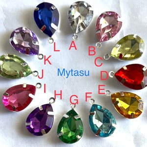 Può includere: Dodici strass di vetro a forma di goccia con montature e anelli argentati. Gli strass sono di vari colori, tra cui verde, blu, rosso, viola, rosa e giallo.