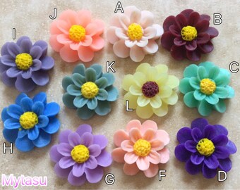 Könnte beinhalten: Zwölf Blumenverzierung aus Harz in verschiedenen Farben, darunter Rosa, Lila, Blau, Gelb und Grün. Jede Blume hat eine gelbe Mitte und ein mehrschichtiges Blütenblattdesign.