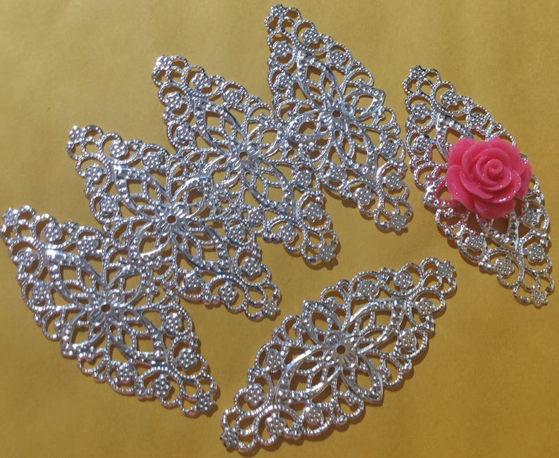 May include: Five silver filigree metal embellishments in the shape of leaves with a pink flower in the center of one of the leaves.