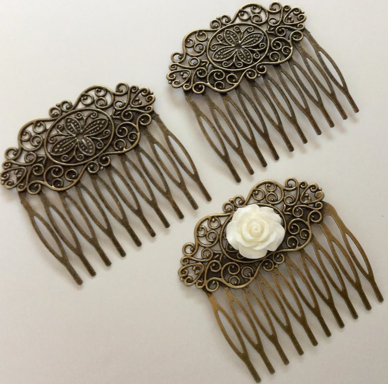 May include: Three antique bronze hair combs with ornate filigree designs. Two combs feature a central oval design, while the third has a white rose accent. The combs have multiple tines for securing in hair.