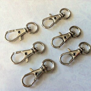 May include: Six silver swivel lobster clasps, also known as trigger snaps, are shown against a white background. These clasps are commonly used for attaching keychains, lanyards, and other accessories.