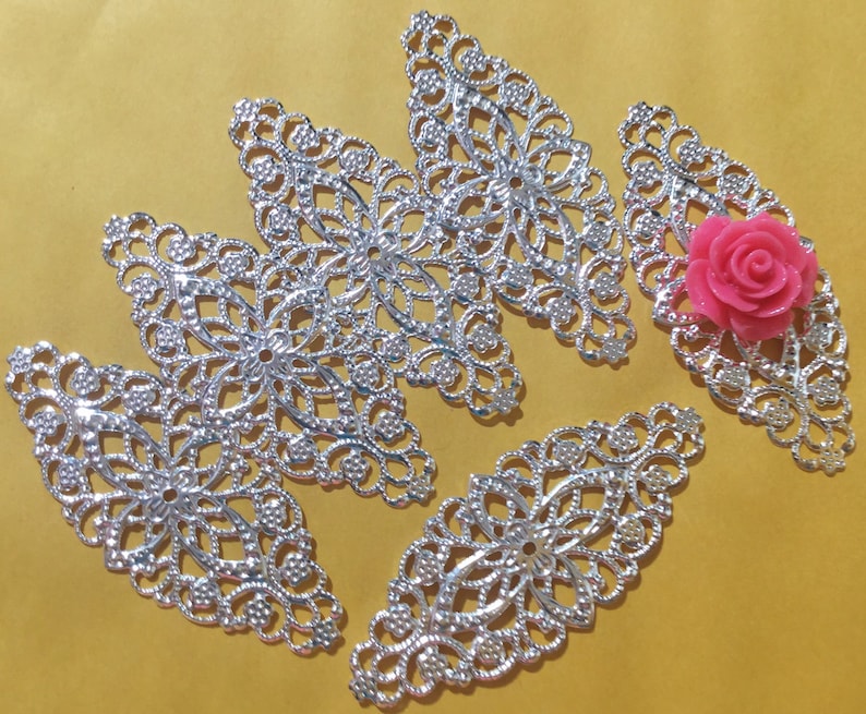 May include: Five silver filigree metal embellishments in the shape of a leaf with a pink flower in the center of one of the embellishments.