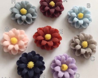 Op de afbeelding: Tien verschillende gekleurde stoffen bloemenversieringen. De bloemen zijn allemaal dezelfde vorm en grootte, met een geel centrum en meerdere lagen bloemblaadjes. De kleuren zijn bordeauxrood, lichtblauw, roze, rood, grijs, donkerblauw, lavendel en lichtblauw.