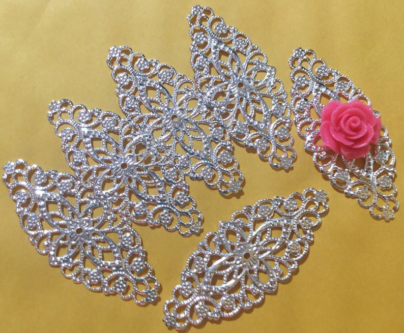 May include: Five silver filigree metal embellishments in the shape of a leaf with a pink flower in the center of one of the embellishments.