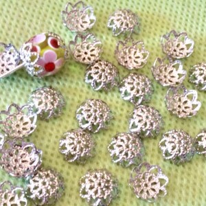 May include: A close-up of a group of silver filigree bead caps. The caps are arranged in a random pattern on a green surface.