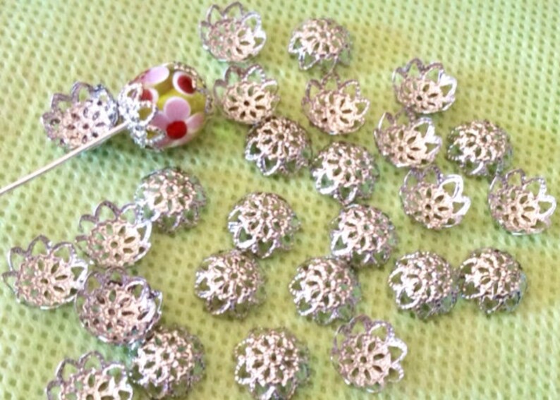 May include: A close-up of a group of silver filigree bead caps. The caps are arranged in a random pattern on a green surface.