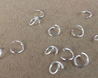 May include: A close-up of a group of silver jump rings, scattered on a brown surface. The jump rings are small and circular, with a gap in the circle.
