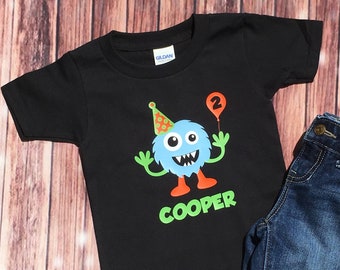 May include: Black t-shirt with a blue monster wearing a party hat and holding a balloon with the number 2. The text "COOPER" is printed below the monster.