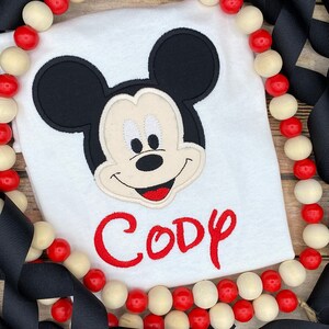 Puede incluir: Camiseta blanca con una cabeza de Mickey Mouse bordada en rojo y el nombre "Cody" en letras bordadas rojas.