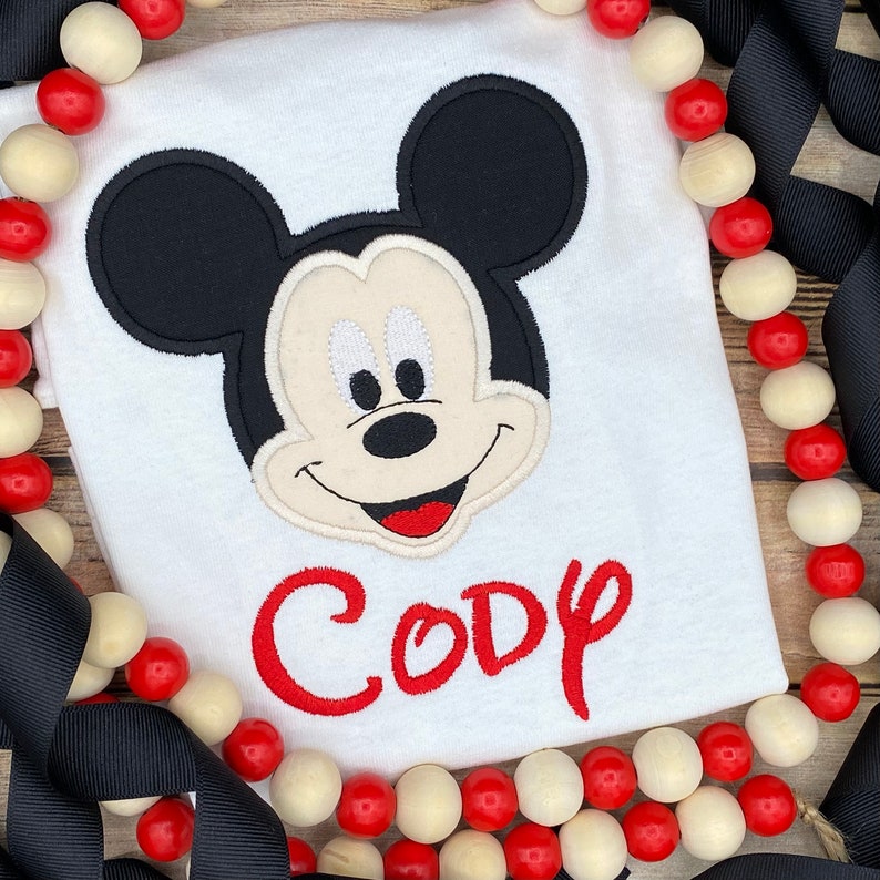 Puede incluir: Camiseta blanca con una cabeza de Mickey Mouse bordada en rojo y el nombre "Cody" en letras bordadas rojas.