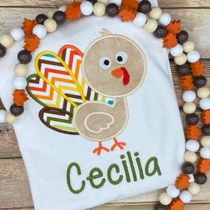 May include: White t-shirt featuring an embroidered turkey design with a colorful chevron tail. The turkey is tan with orange feet and a red wattle. The name "Cecilia" is embroidered in green script below the turkey. A beaded garland with wooden leaves frames the shirt.