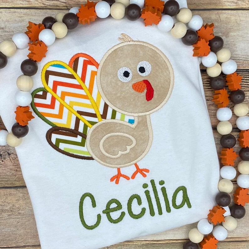 May include: White t-shirt featuring an embroidered turkey design with a colorful chevron tail. The turkey is tan with orange feet and a red wattle. The name "Cecilia" is embroidered in green script below the turkey. A beaded garland with wooden leaves frames the shirt.