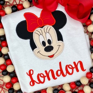 May include: White t-shirt with a red and black embroidered Minnie Mouse face and the name "London" in red embroidery.