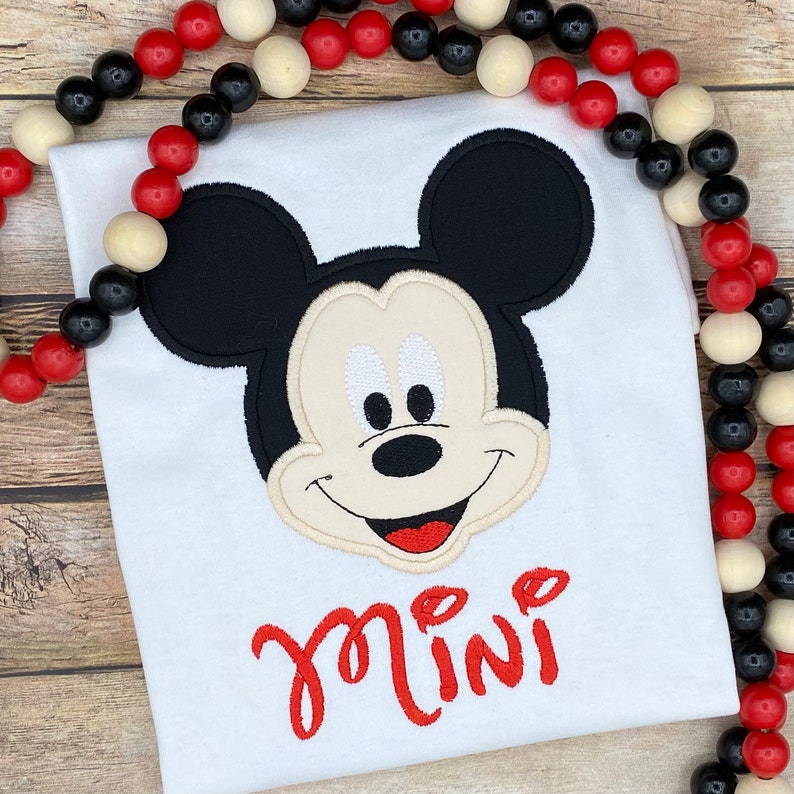 Puede incluir: Camiseta blanca con una imagen bordada de la cara de Mickey Mouse y la palabra "mini" en rojo.