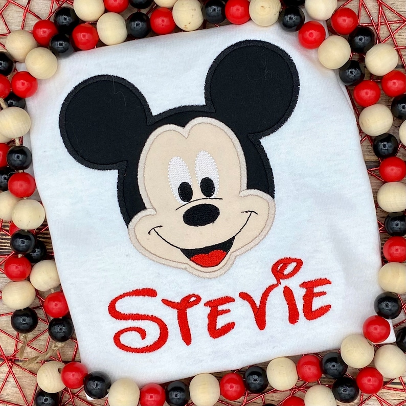 Puede incluir: Camiseta blanca con una cabeza de Mickey Mouse bordada en rojo y el nombre "Stevie" en bordado rojo.
