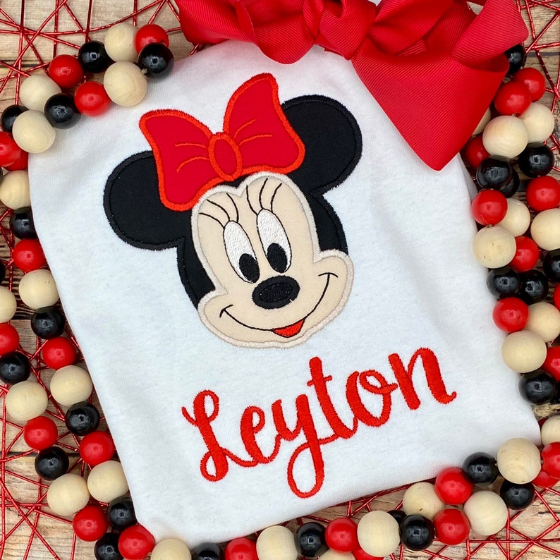 May include: White t-shirt with a red and black embroidered Minnie Mouse head and the name "Leyton" in red embroidery.