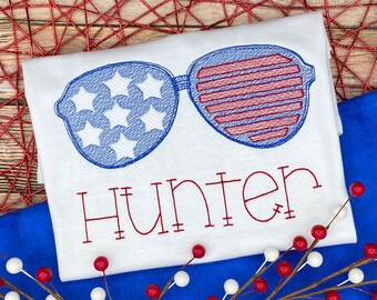 Könnte beinhalten: Weißes T-Shirt mit roten und blauen gestickten Sonnenbrillen und dem Namen "Hunter" in roter Stickerei.