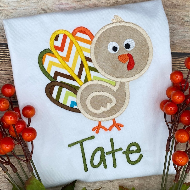 May include: White t-shirt featuring an embroidered turkey design with a beige body, colorful tail feathers, and the name "Tate" in green. The turkey has a red wattle and orange beak. Perfect for Thanksgiving or fall celebrations.