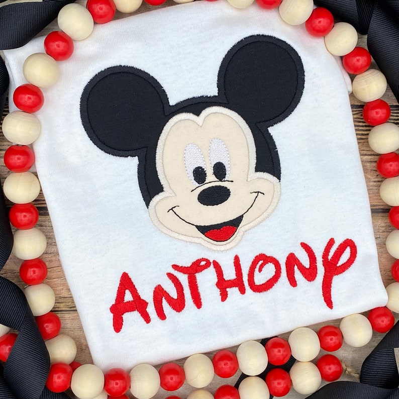 Puede incluir: Camiseta blanca con una cabeza de Mickey Mouse bordada y el nombre "ANTHONY" en rojo.