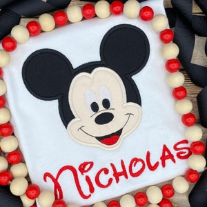 Puede incluir: Camiseta blanca con un rostro de Mickey Mouse bordado y el nombre "Nicholas" en rojo.