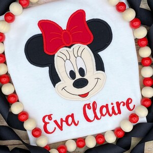 May include: White t-shirt with a red and black embroidered Minnie Mouse design and the name "Eva Claire" in red embroidery.