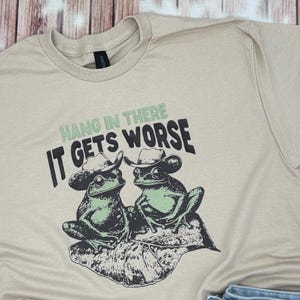 May include: Beige t-shirt featuring a graphic of two frogs wearing cowboy hats, sitting on a leaf. The text "Hang in there, it gets worse" is printed above the frogs in green and black.