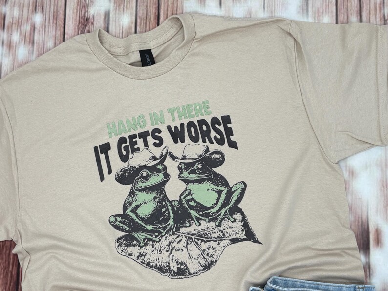 May include: Beige t-shirt featuring a graphic of two frogs wearing cowboy hats, sitting on a leaf. The text "Hang in there, it gets worse" is printed above the frogs in green and black.