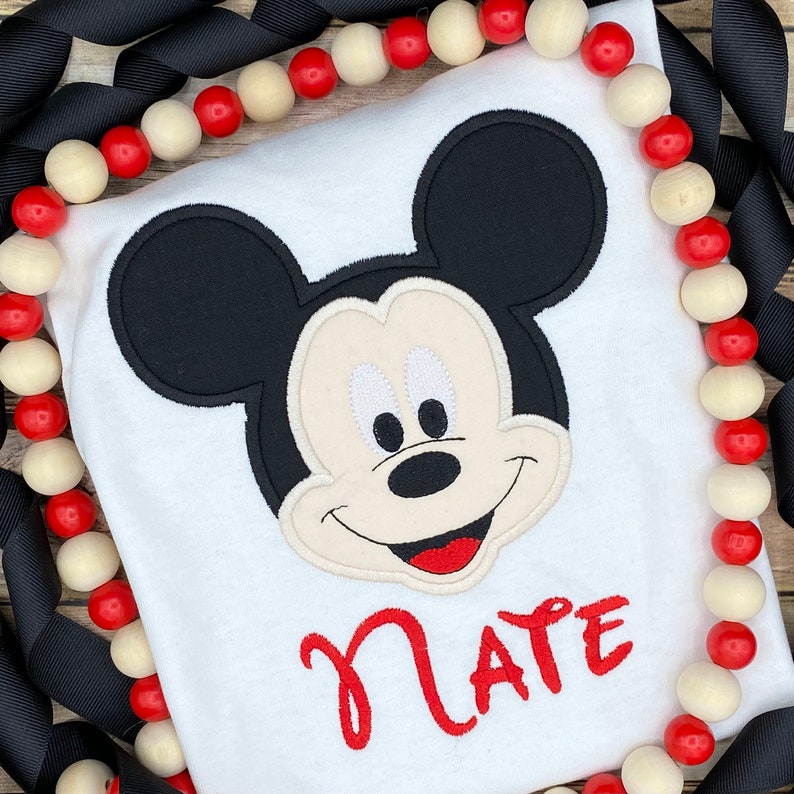 Puede incluir: Camiseta blanca con una cabeza de Mickey Mouse bordada y el nombre "Nate" en rojo.